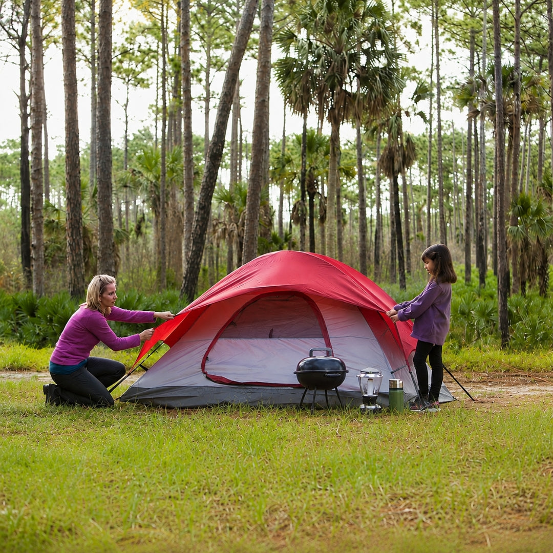 Two people setting up a red tent in a forested area.