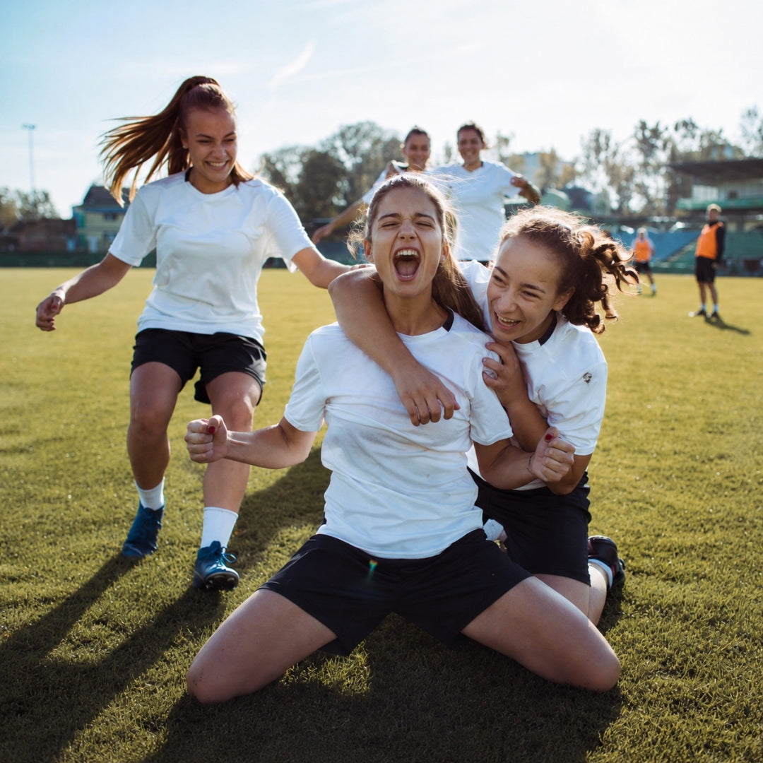 Two female soccer players celebrating on a field with teammates in the background.